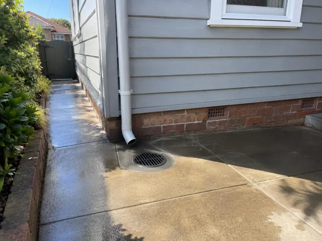 Outdoor grated drain beside a house wall and paved path, with downpipe and brick base in Five Dock
