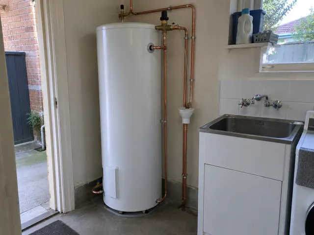 White hot water cylinder with copper pipework beside a laundry sink and tiled wall in a home in Drummoyne